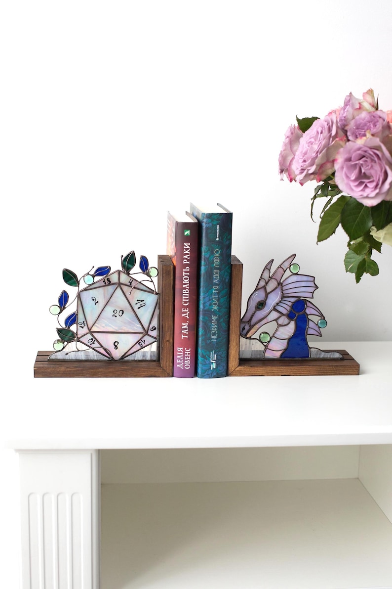 May include: Stained glass bookends with a D20 die and a dragon design. The bookends are made of wood and hold two books. The die is white with black numbers, and the dragon is purple and blue. A bouquet of pink roses is in the background.