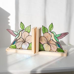 Pu&ograve; includere: Reggilibri in legno con un design a vetrata raffigurante colibr&igrave; e fiori. I reggilibri hanno una base in legno chiaro e tengono i libri tra due supporti in legno. Il colibr&igrave; ha ali verdi, viola e blu, con un fiore rosa.