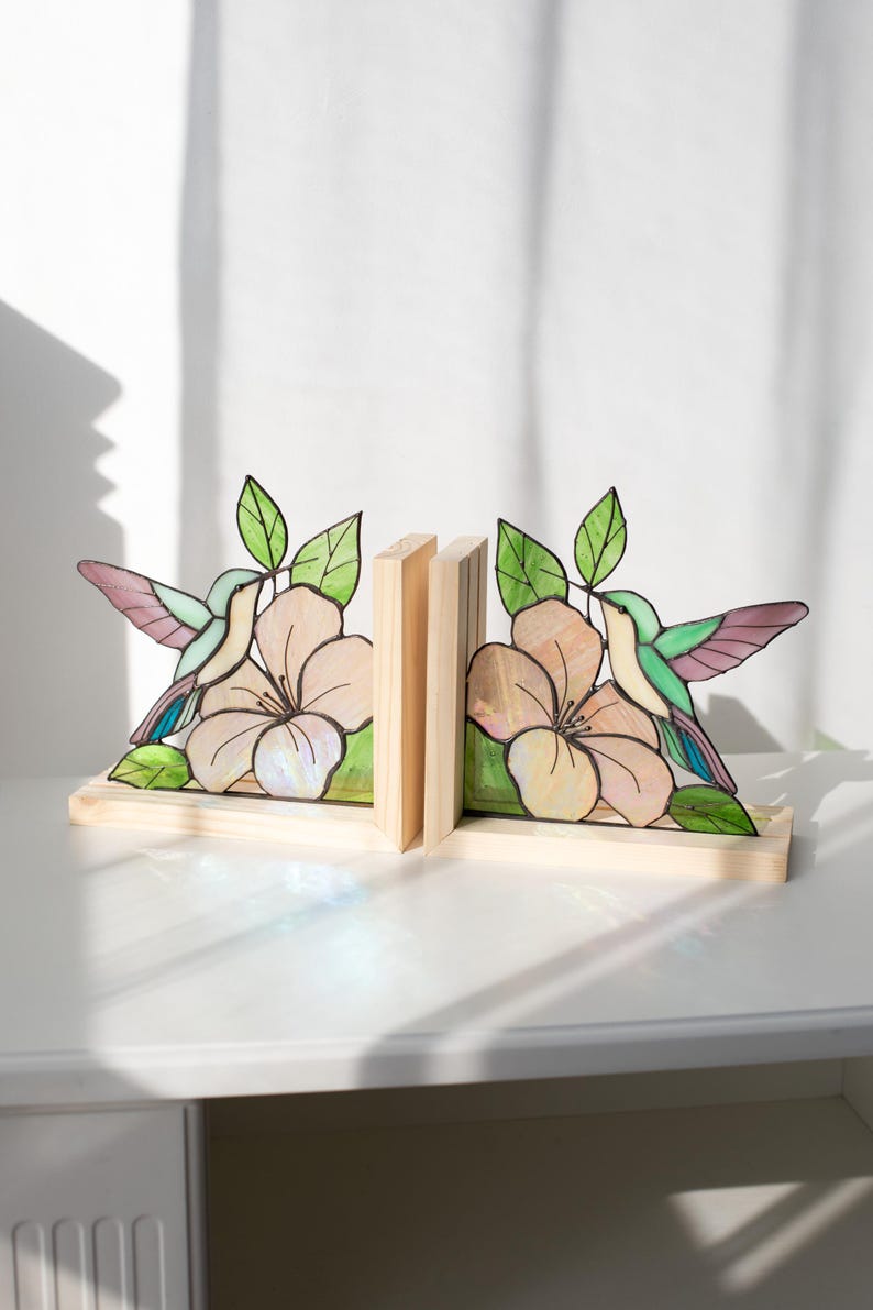 Pu&ograve; includere: Reggilibri in legno con un design a vetrata raffigurante colibr&igrave; e fiori. I reggilibri hanno una base in legno chiaro e tengono i libri tra due supporti in legno. Il colibr&igrave; ha ali verdi, viola e blu, con un fiore rosa.
