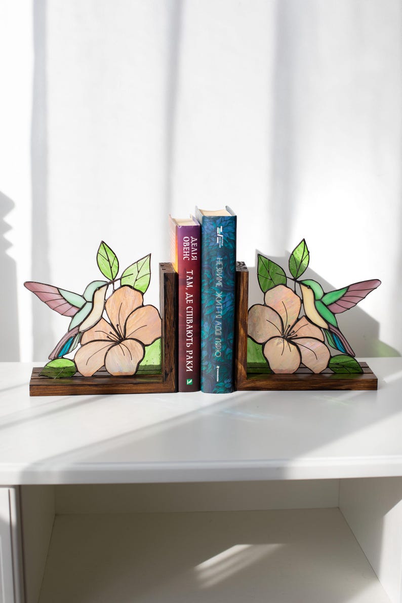 Pu&ograve; includere: Reggilibri con colibr&igrave; e fiori in vetro colorato. I reggilibri sono in legno e presentano un colorato design di colibr&igrave; con accenti rosa, verdi e viola. I reggilibri sostengono tre libri.