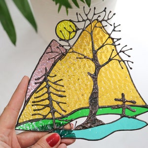 May include: A stained glass window hanging features a yellow sun, a pink mountain, a green field, a blue lake, and a bare tree with brown branches.