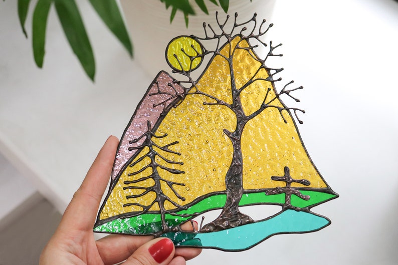 May include: A stained glass window hanging features a yellow sun, a pink mountain, a green field, a blue lake, and a bare tree with brown branches.