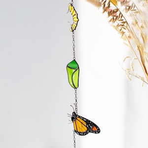 May include: A stained glass mobile featuring a monarch butterfly life cycle. The mobile includes a yellow egg, a green chrysalis, and two orange and black butterflies, all suspended by a chain. The artwork is designed to hang as a decorative piece.