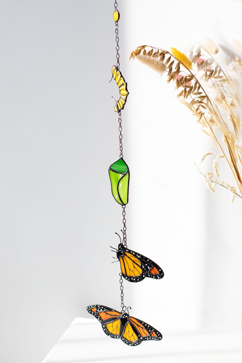 May include: A stained glass mobile featuring a monarch butterfly life cycle. The mobile includes a yellow egg, a green chrysalis, and two orange and black butterflies, all suspended by a chain. The artwork is designed to hang as a decorative piece.
