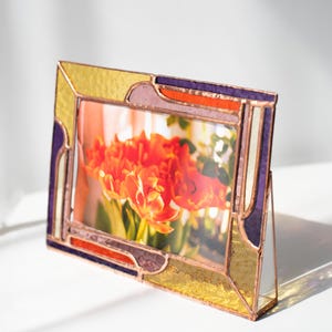 May include: A stained glass picture frame with a colorful design. The frame features gold, purple, orange, and white glass panels. The frame is angled, displaying a photo of orange flowers.