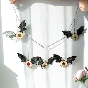 May include: A hanging mobile featuring five stained glass bat figures, each with a central realistic eyeball design. The bats have black wings and irises in various colours including green, red, blue, and brown, all connected by thin metal chains.