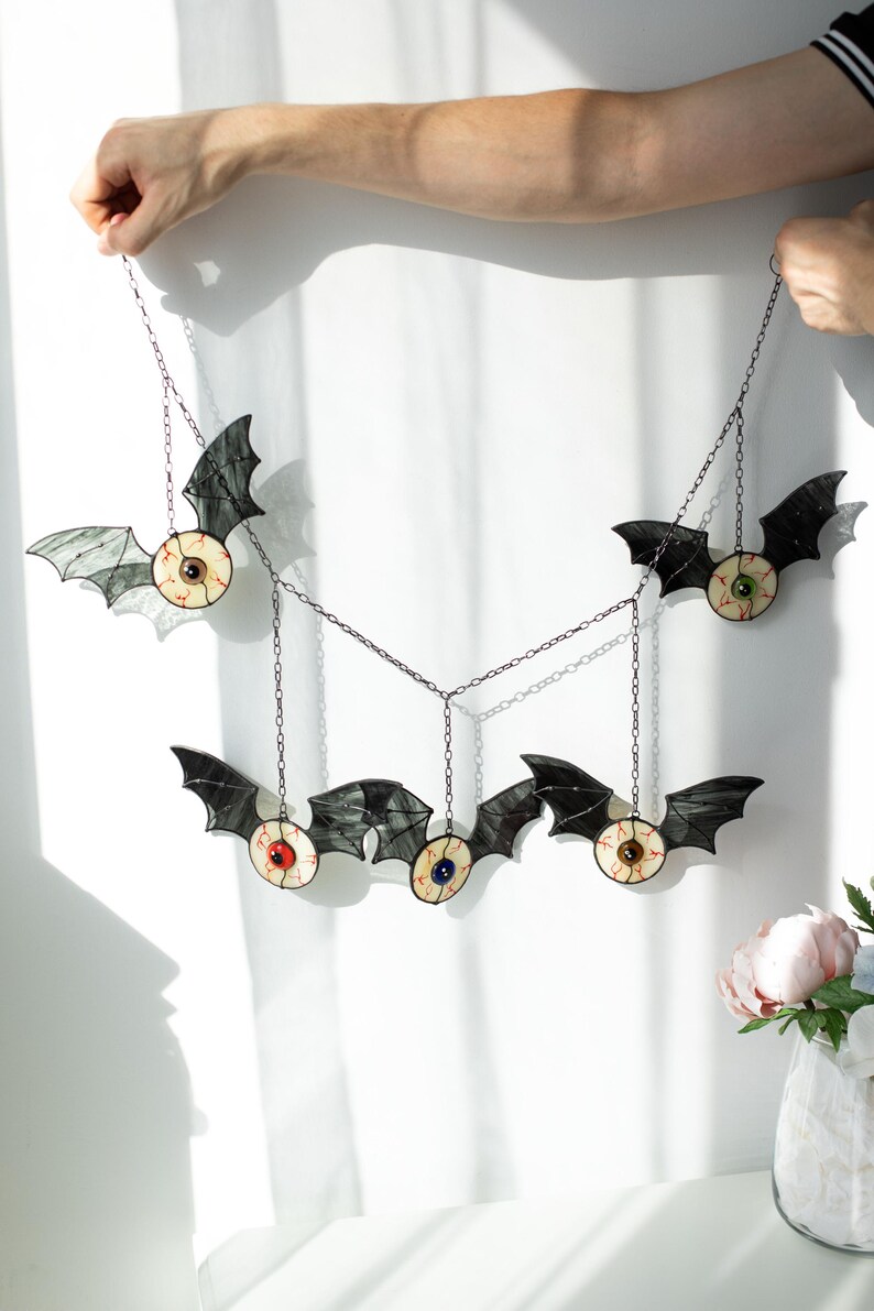 May include: A hanging mobile featuring five stained glass bat figures, each with a central realistic eyeball design. The bats have black wings and irises in various colours including green, red, blue, and brown, all connected by thin metal chains.