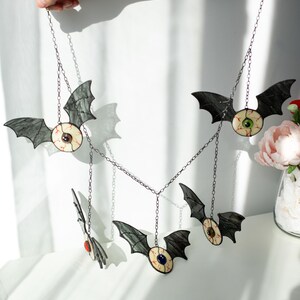 May include: A hanging mobile featuring five stained glass bat figures, each with a central eyeball design. The bats have dark grey wings and cream-coloured bodies with red vein details. Each eyeball has a different coloured iris, suspended by chains.