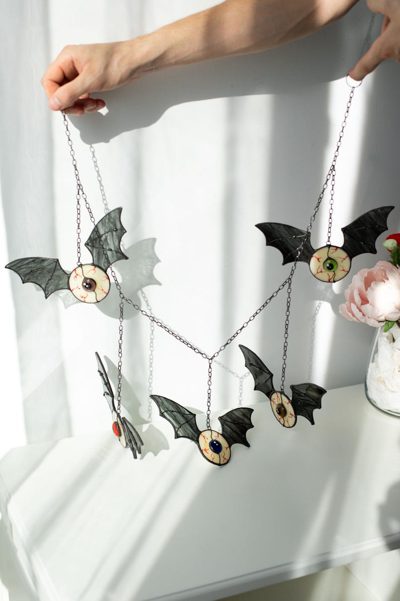 May include: A hanging mobile featuring five stained glass bat figures, each with a central eyeball design. The bats have dark grey wings and cream-coloured bodies with red vein details. Each eyeball has a different coloured iris, suspended by chains.