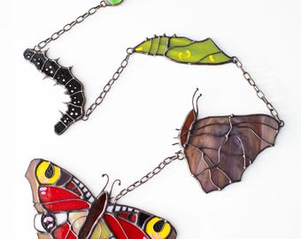 Puede incluir: Un móvil de mariposa de vidrio coloreado con una oruga negra, una crisálida verde y dos mariposas. Las mariposas son marrones y rojas con detalles amarillos.