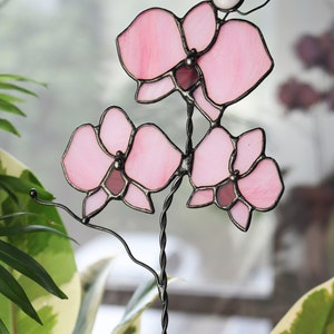 K&ouml;nnte beinhalten: Eine rosa gef&auml;rbte Glasorchidee mit einem schwarzen Metallstiel. Die Orchidee besteht aus mehreren Glasst&uuml;cken, die mit schwarzem Metall verbunden sind.