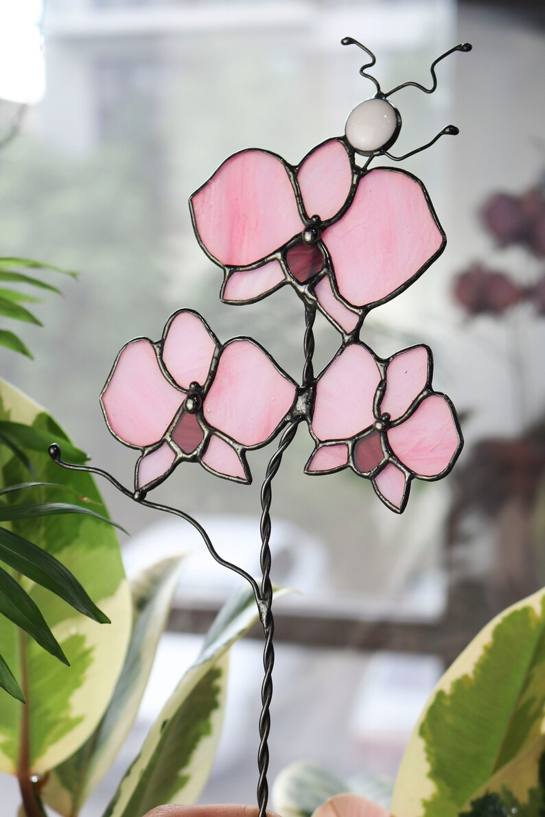 K&ouml;nnte beinhalten: Eine rosa gef&auml;rbte Glasorchidee mit einem schwarzen Metallstiel. Die Orchidee besteht aus mehreren Glasst&uuml;cken, die mit schwarzem Metall verbunden sind.
