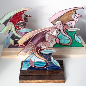 Op de afbeelding: Gebrandschilderde glazen drakenbeelden in tinten paars, roze en rood, op houten sokkels. De draken hebben uitgespreide vleugels. De sculpturen staan tegen een witte achtergrond. Elke draak is een uniek kunstwerk.