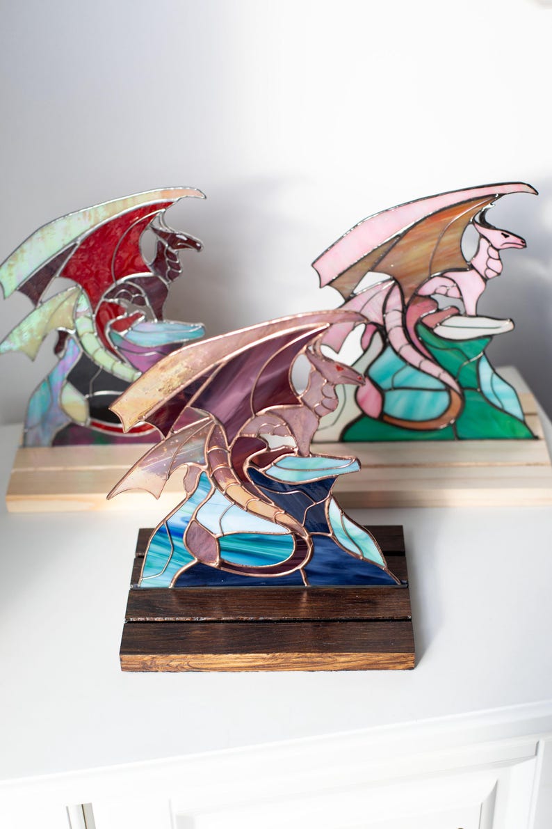 Op de afbeelding: Gebrandschilderde glazen drakenbeelden in tinten paars, roze en rood, op houten sokkels. De draken hebben uitgespreide vleugels. De sculpturen staan tegen een witte achtergrond. Elke draak is een uniek kunstwerk.