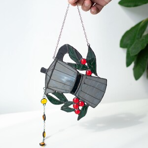 Puede incluir: Un atrapasue&ntilde;os de vitral con forma de cafetera moka, que presenta paneles de vidrio gris transl&uacute;cido, hojas de vidrio verde oscuro y bayas de vidrio rojo brillante. Una peque&ntilde;a cadena con cuentas de vidrio color &aacute;mbar cuelga del lateral.