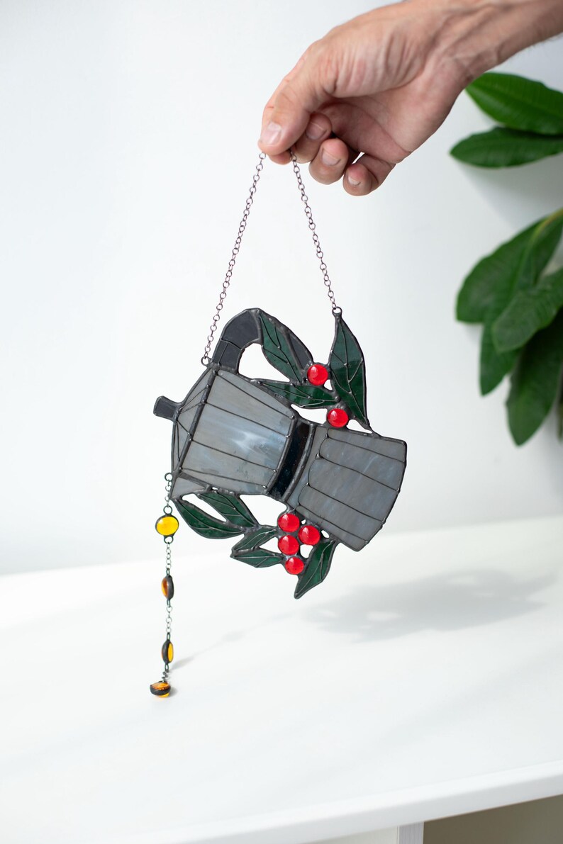 Puede incluir: Un atrapasue&ntilde;os de vitral con forma de cafetera moka, que presenta paneles de vidrio gris transl&uacute;cido, hojas de vidrio verde oscuro y bayas de vidrio rojo brillante. Una peque&ntilde;a cadena con cuentas de vidrio color &aacute;mbar cuelga del lateral.