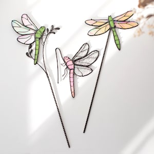 Può includere: Tre ornamenti a forma di libellula in vetro colorato con steli in metallo. Una libellula è verde con ali rosa chiaro, una è rosa con ali trasparenti e una è verde con ali color arancio. Presentano saldature in metallo scuro e steli sottili.