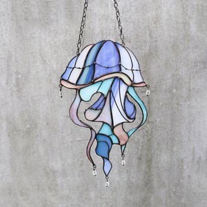 Op de afbeelding: Een gebrandschilderd glas-zonnevanger in de vorm van een kwal met blauwe, paarse en roze tinten. De kwal heeft een klokvormige bovenkant en lange, vloeiende tentakels. De zonnevanger hangt aan een ketting.