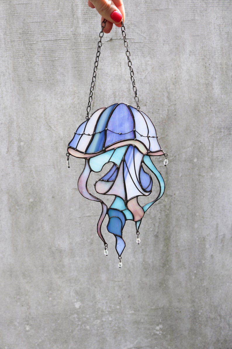 Op de afbeelding: Een gebrandschilderd glas-zonnevanger in de vorm van een kwal met blauwe, paarse en roze tinten. De kwal heeft een klokvormige bovenkant en lange, vloeiende tentakels. De zonnevanger hangt aan een ketting.