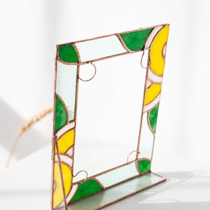 May include: A stained glass picture frame with a copper-colored metal frame. The frame features sections of green, yellow, and white glass. The frame is designed to stand upright on a flat surface. The frame is a decorative item.