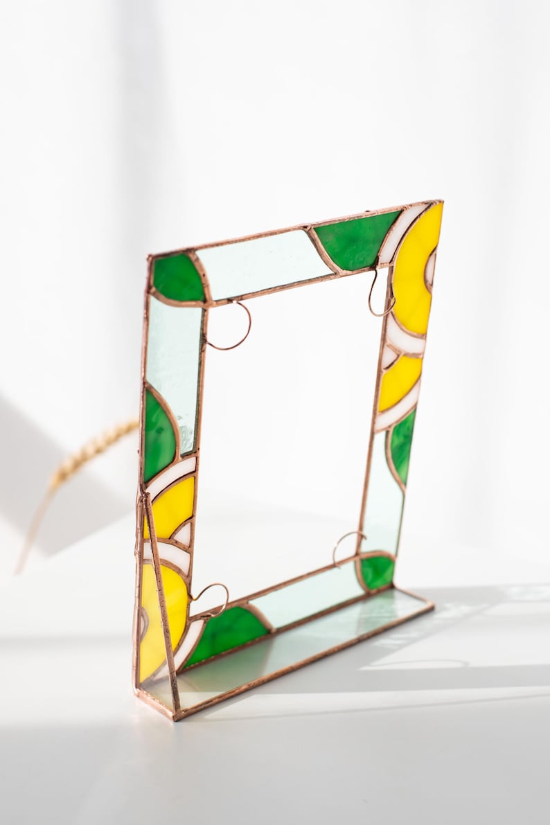 May include: A stained glass picture frame with a copper-colored metal frame. The frame features sections of green, yellow, and white glass. The frame is designed to stand upright on a flat surface. The frame is a decorative item.