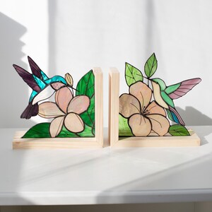 Pu&ograve; includere: Un paio di fermalibri in legno con disegni di colibr&igrave; e fiori in vetro colorato. I colibr&igrave; sono turchesi, viola e bianchi. I fiori sono rosa chiaro con foglie verdi. Su una superficie bianca.