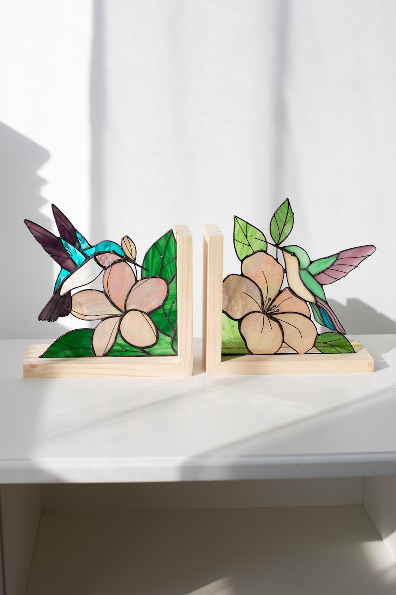 Pu&ograve; includere: Un paio di fermalibri in legno con disegni di colibr&igrave; e fiori in vetro colorato. I colibr&igrave; sono turchesi, viola e bianchi. I fiori sono rosa chiaro con foglie verdi. Su una superficie bianca.
