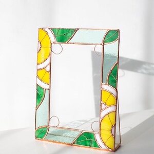 May include: A stained glass picture frame with a vibrant design. The frame features yellow, green, and white geometric patterns, with copper-colored borders. The frame is standing upright, showcasing its decorative elements.