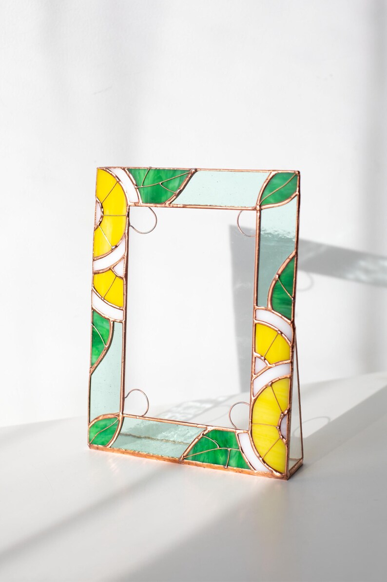 May include: A stained glass picture frame with a vibrant design. The frame features yellow, green, and white geometric patterns, with copper-colored borders. The frame is standing upright, showcasing its decorative elements.