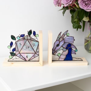 May include: Two stained glass bookends. One is a D20 die with numbers, the other a dragon head. Both are set in wooden frames. The die has green and blue leaves. The dragon is purple and blue. A bouquet of purple roses is in the background.