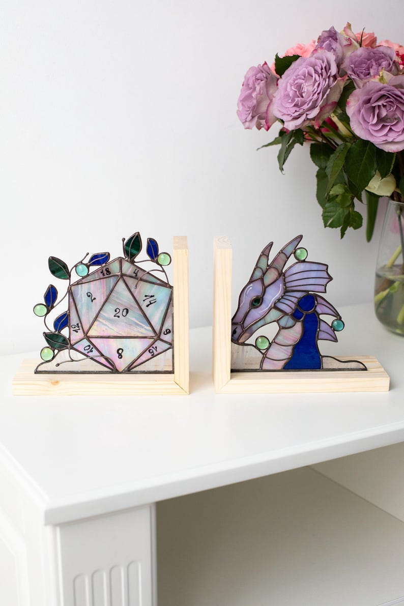 May include: Two stained glass bookends. One is a D20 die with numbers, the other a dragon head. Both are set in wooden frames. The die has green and blue leaves. The dragon is purple and blue. A bouquet of purple roses is in the background.