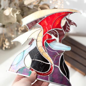 Op de afbeelding: Gebrandschilderde glazen drakenbeeld met rode, witte en paarse vleugels. De draak heeft een gele en rode romp en staat tegen een achtergrond van zwart, paars en iriserend glas. Het kunstwerk wordt in een hand gehouden.