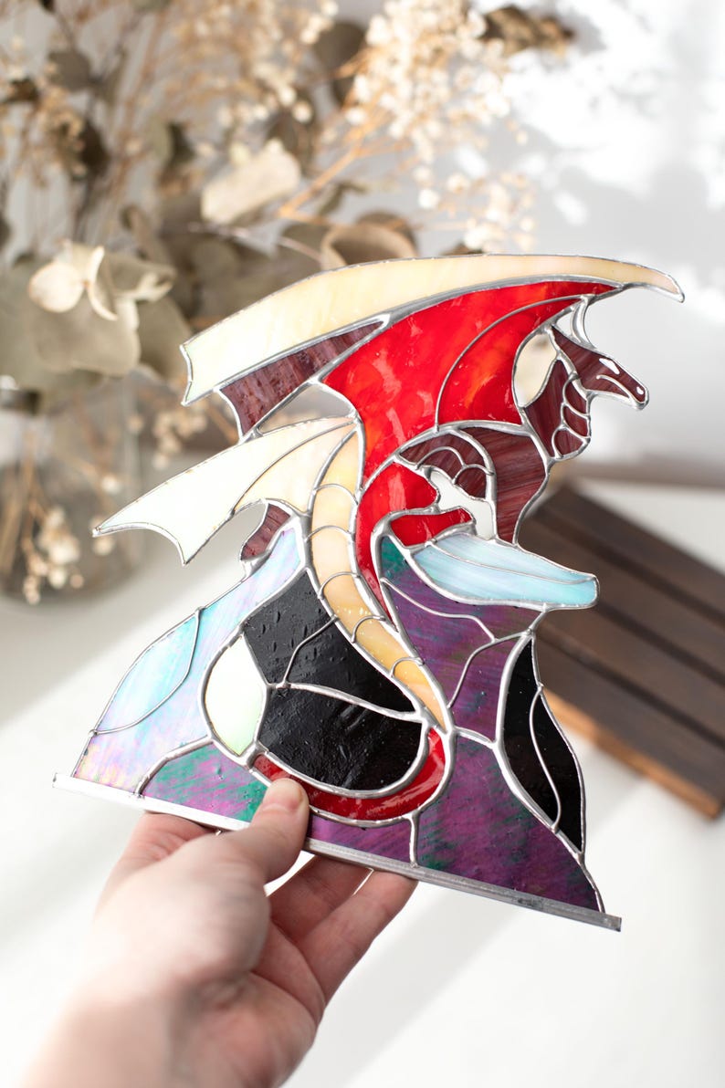 Op de afbeelding: Gebrandschilderde glazen drakenbeeld met rode, witte en paarse vleugels. De draak heeft een gele en rode romp en staat tegen een achtergrond van zwart, paars en iriserend glas. Het kunstwerk wordt in een hand gehouden.