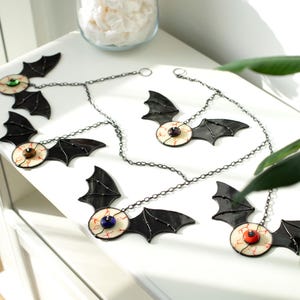 May include: A set of four hanging ornaments featuring black stained glass bat wings attached to a central eyeball design. Each eyeball has a white sclera with red veins and a coloured iris in blue, brown, orange, or purple, suspended by metal chains.