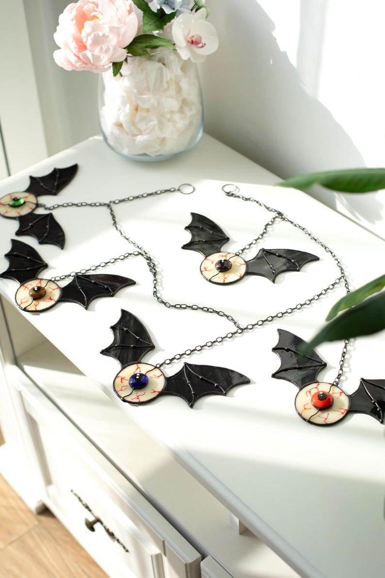 May include: A set of four hanging ornaments featuring black stained glass bat wings attached to a central eyeball design. Each eyeball has a white sclera with red veins and a coloured iris in blue, brown, orange, or purple, suspended by metal chains.