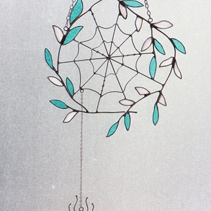 May include: A stained glass spider web suncatcher with teal and white leaves hanging from a silver chain. A small spider with an orange body hangs from the bottom of the chain.