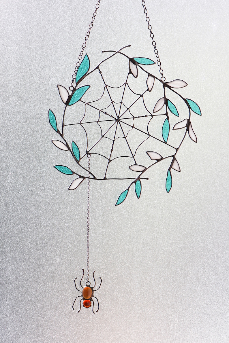 May include: A stained glass spider web suncatcher with teal and white leaves hanging from a silver chain. A small spider with an orange body hangs from the bottom of the chain.
