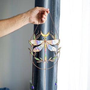 May include: A stained glass dragonfly suncatcher with iridescent wings and a crescent moon. The dragonfly is surrounded by green leaves and hangs from a copper chain with purple and green beads.