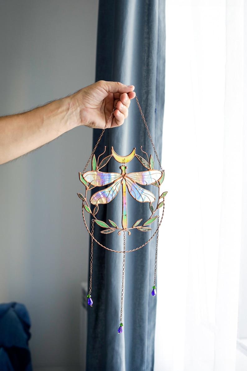 May include: A stained glass dragonfly suncatcher with iridescent wings and a crescent moon. The dragonfly is surrounded by green leaves and hangs from a copper chain with purple and green beads.