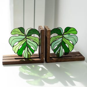 K&ouml;nnte beinhalten: Zwei Buchst&uuml;tzen aus Holz mit Monstera-Blatt-Designs aus Glasmalerei. Die Bl&auml;tter sind in verschiedenen Gr&uuml;nt&ouml;nen gehalten und in einem dunklen Rahmen eingefasst. Die Buchst&uuml;tzen stehen auf einer wei&szlig;en Oberfl&auml;che.