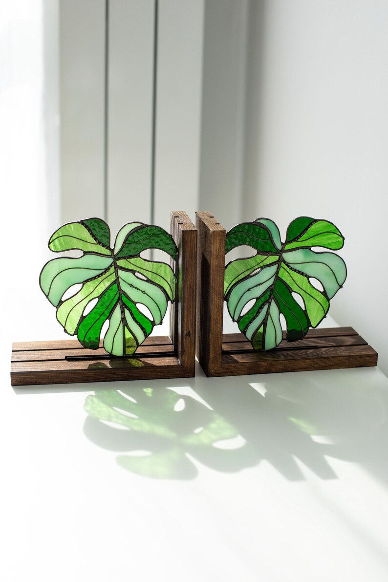 K&ouml;nnte beinhalten: Zwei Buchst&uuml;tzen aus Holz mit Monstera-Blatt-Designs aus Glasmalerei. Die Bl&auml;tter sind in verschiedenen Gr&uuml;nt&ouml;nen gehalten und in einem dunklen Rahmen eingefasst. Die Buchst&uuml;tzen stehen auf einer wei&szlig;en Oberfl&auml;che.