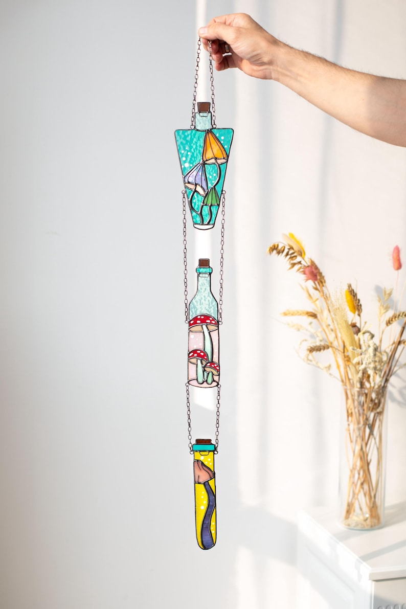 May include: Three stained glass bottles hanging from a chain. The bottles are filled with different colored mushrooms. The top bottle is teal, the middle bottle is clear with pink and red mushrooms, and the bottom bottle is yellow with purple mushrooms.