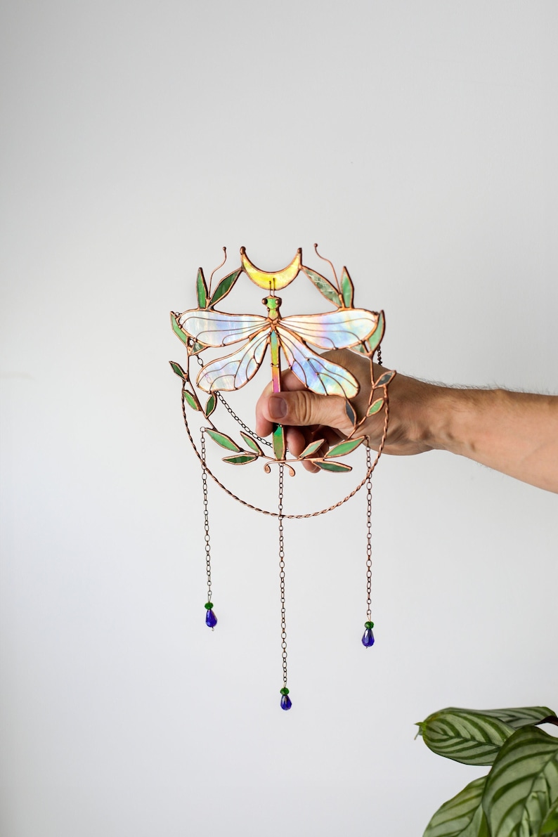May include: A stained glass dragonfly with iridescent wings, surrounded by a copper wire wreath with green leaves and a crescent moon. Three chains with blue and green glass beads hang from the bottom.
