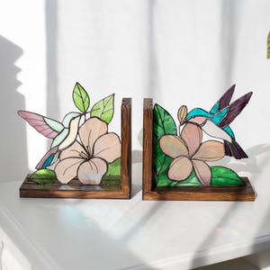 Pu&ograve; includere: Un paio di fermalibri in legno con disegni di colibr&igrave; e fiori in vetro colorato. I colibr&igrave; hanno ali rosa, blu e viola. I fiori sono rosa chiaro con foglie verdi. I fermalibri sono su una superficie bianca.