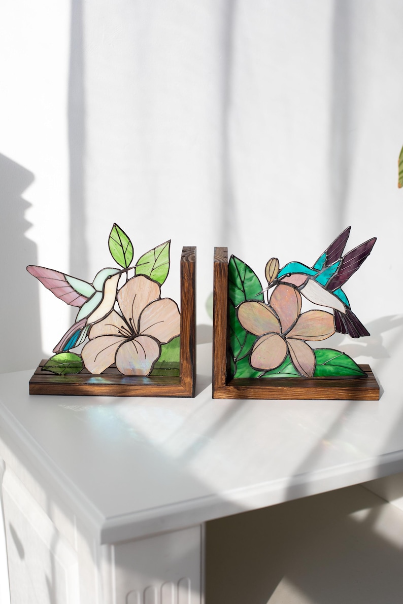 Pu&ograve; includere: Un paio di fermalibri in legno con disegni di colibr&igrave; e fiori in vetro colorato. I colibr&igrave; hanno ali rosa, blu e viola. I fiori sono rosa chiaro con foglie verdi. I fermalibri sono su una superficie bianca.
