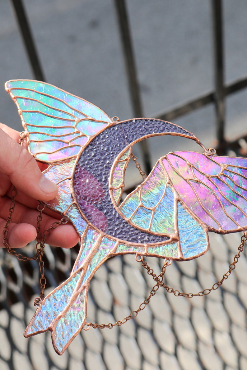 May include: A stained glass suncatcher featuring a crescent moon and iridescent butterfly wings. The suncatcher is suspended from a copper chain.