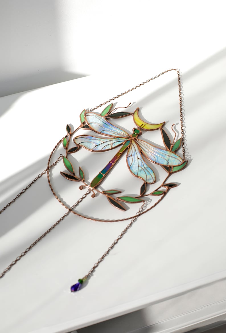 May include: A stained glass dragonfly suncatcher with iridescent wings, a crescent moon, and green leaves. The suncatcher hangs from a copper chain with a purple crystal bead.