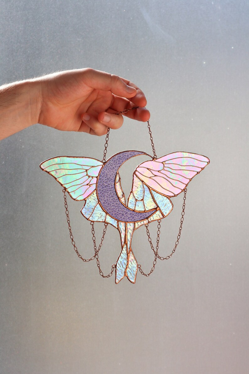 May include: A stained glass moon and moth suncatcher with iridescent wings and a purple moon. The suncatcher is hanging from a copper chain.