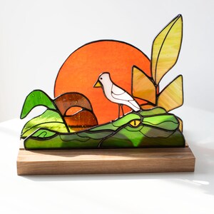 Op de afbeelding: Gebrandschilderde glazen sculptuur van een alligator met een witte vogel op zijn rug. De sculptuur heeft groen, geel en oranje glas, geplaatst op een houten voet. Een feloranje zon is op de achtergrond.