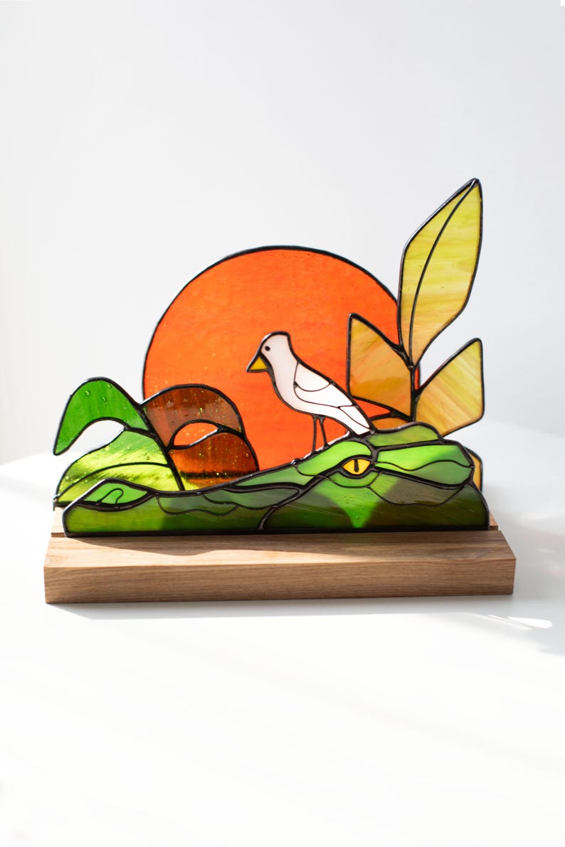 Op de afbeelding: Gebrandschilderde glazen sculptuur van een alligator met een witte vogel op zijn rug. De sculptuur heeft groen, geel en oranje glas, geplaatst op een houten voet. Een feloranje zon is op de achtergrond.
